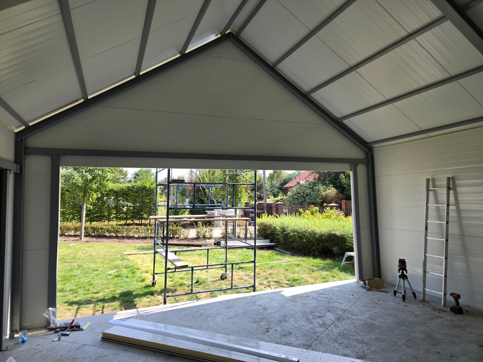 Wnętrze garażu w trakcie montażu z widocznym rusztowaniem, drabiną i narzędziami na betonowej posadzce, z otwartym widokiem na zielony ogród.