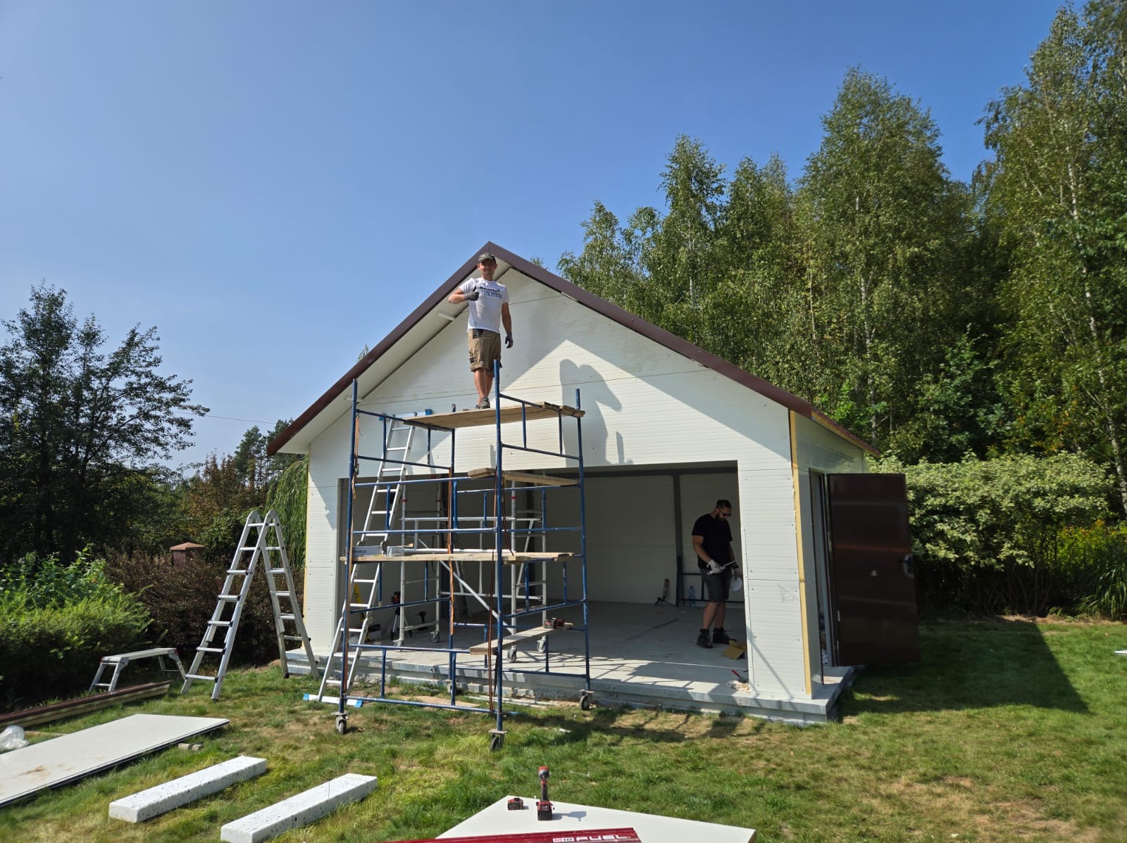 Montaż białej bramy garażowej w Siensku, widok z zewnątrz: dwaj monterzy na rusztowaniu i wewnątrz, elementy konstrukcyjne leżą na trawie, częściowo otwarte drzwi wejściowe.