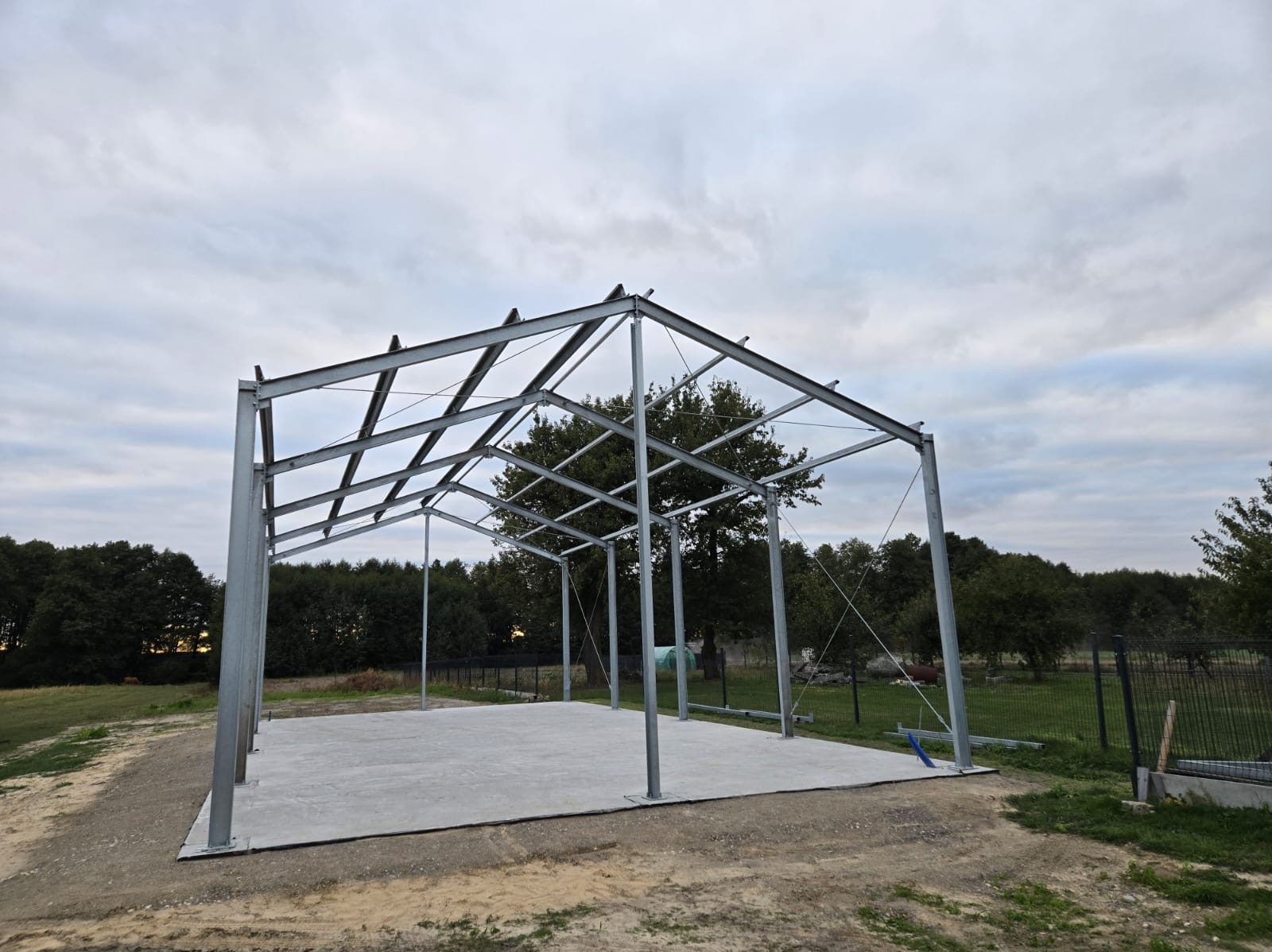 Stalowa konstrukcja szkieletowa budynku gospodarczego na betonowej płycie, widok zewnętrzny w pochmurny dzień.