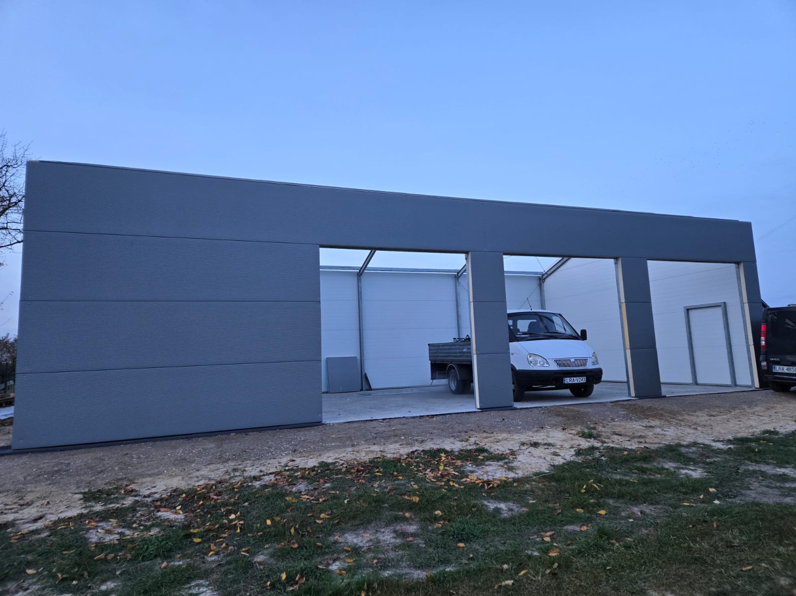 Nowoczesny, szary garaż z przesuwną częścią i dwoma otwartymi wjazdami, wewnątrz zaparkowany biały samochód dostawczy.