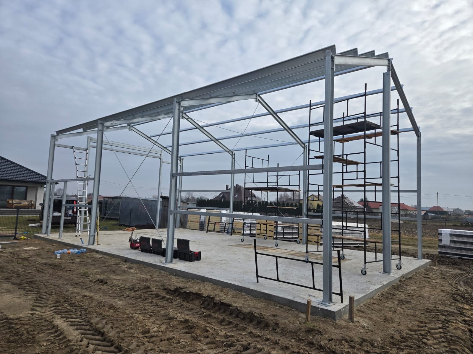 Konstrukcja stalowa szkieletu budynku w trakcie montażu, widoczne rusztowania, drabina i narzędzia na betonowej wylewce, ziemia wokół.