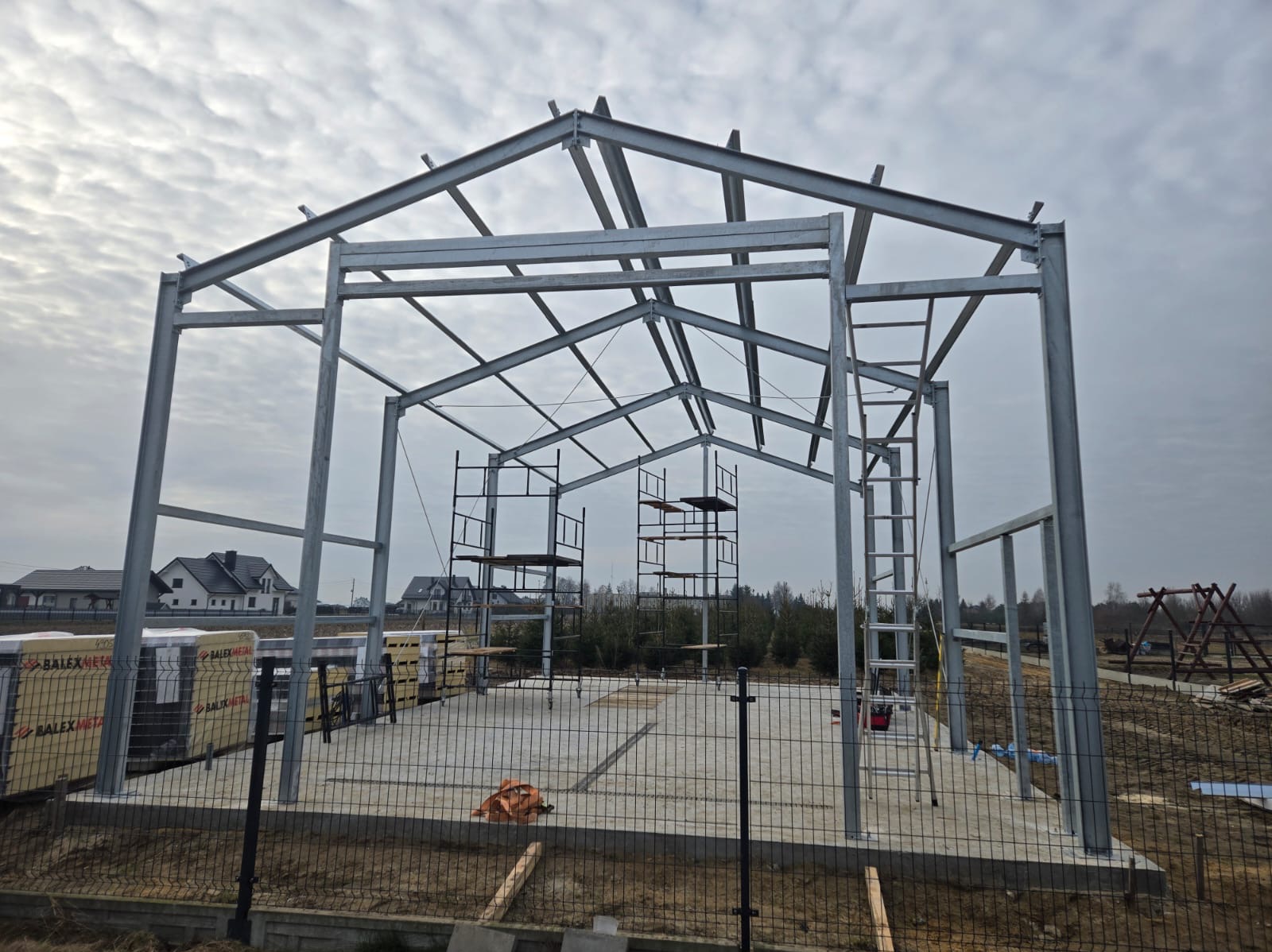 Stalowa konstrukcja hali w trakcie montażu na betonowej płycie, widoczne rusztowania i materiały budowlane, w tle domy jednorodzinne.