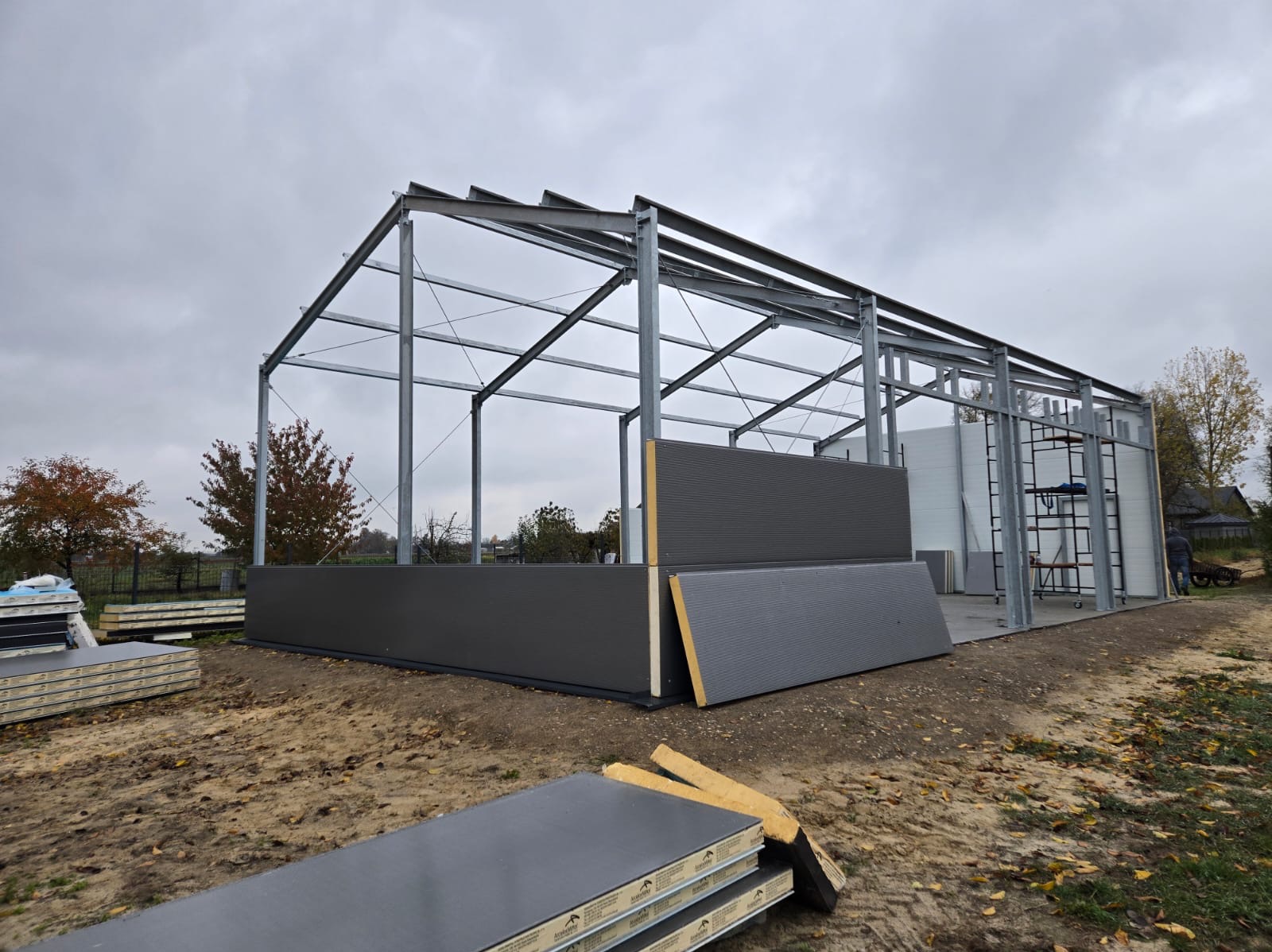 Montaż szkieletu stalowego budynku gospodarczego z częściowo zamontowanymi panelami ściennymi na fundamencie, widoczne składowane panele warstwowe, pochmurne niebo.
