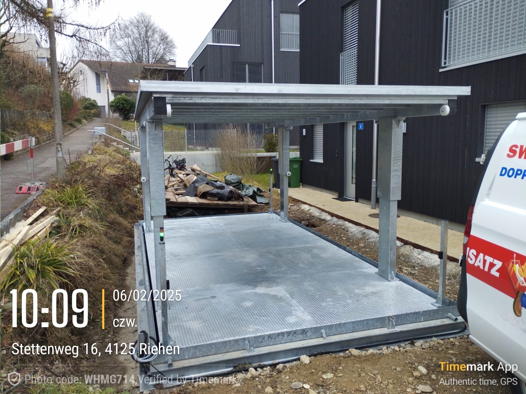 Stalowa konstrukcja platformy podziemnego parkingu dla samochodów w trakcie montażu, widoczna instalacja elektryczna, w tle budynek z ciemnego drewna i teren budowy.