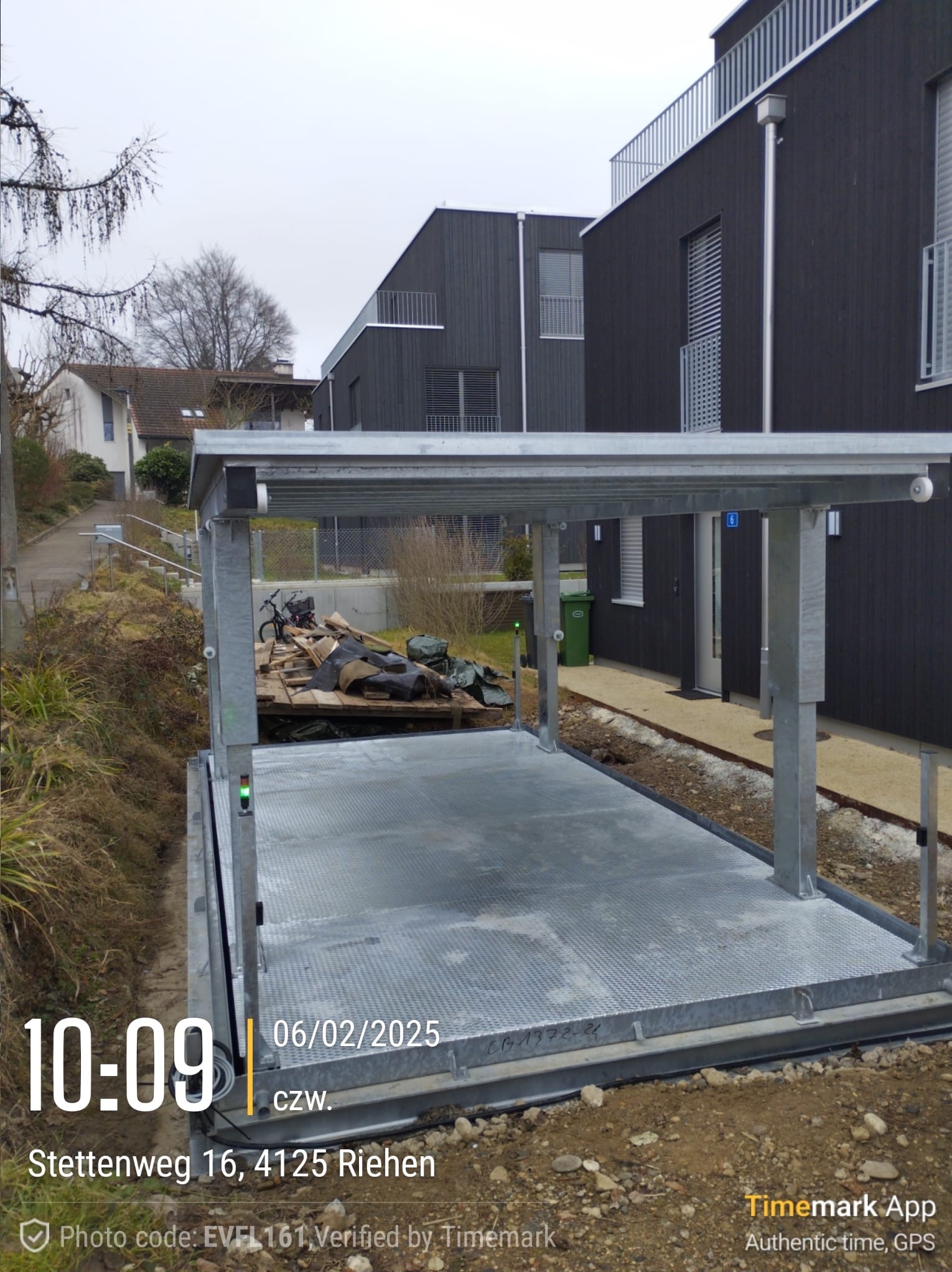 Zewnętrzna platforma podnosząca, prawdopodobnie dla samochodu, z metalową konstrukcją nośną, na tle nowoczesnego budynku mieszkalnego z ciemnego drewna i zielonego kosza na śmieci.