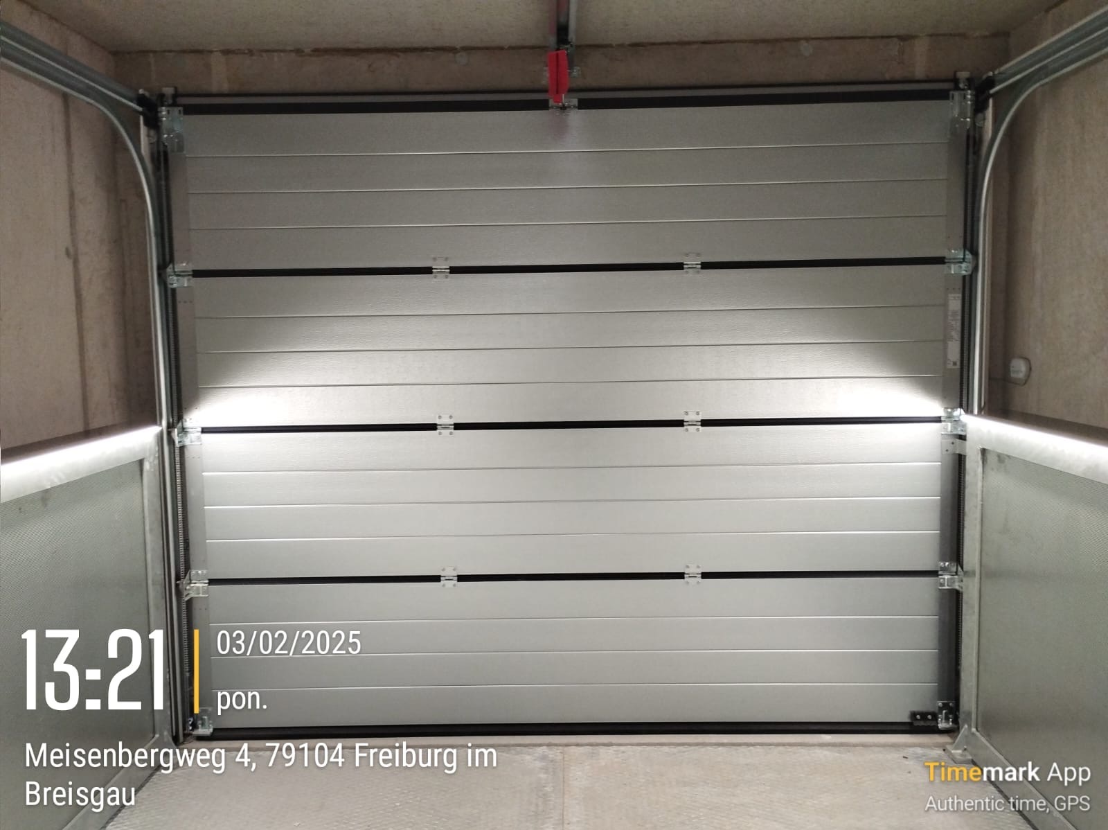 Zamknięta, segmentowa, srebrna brama garażowa z widocznymi zawiasami i prowadnicami, sfotografowana od wewnątrz garażu. Na dole zdjęcia widoczny jest znak wodny z datą i godziną.