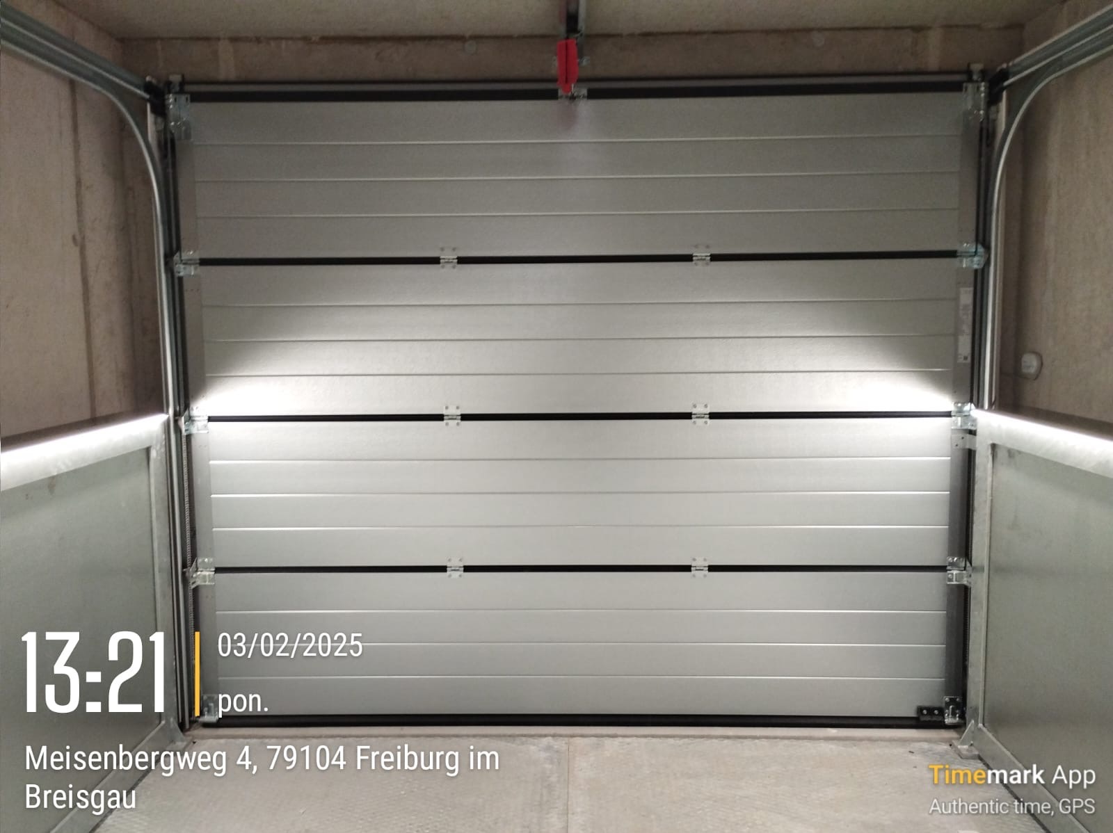 Zamknięta, segmentowa brama garażowa w kolorze jasnoszarym, widok od wewnątrz garażu, z widocznymi elementami mocującymi i prowadnicami, z datą i godziną wykonania zdjęcia nałożoną na obraz.