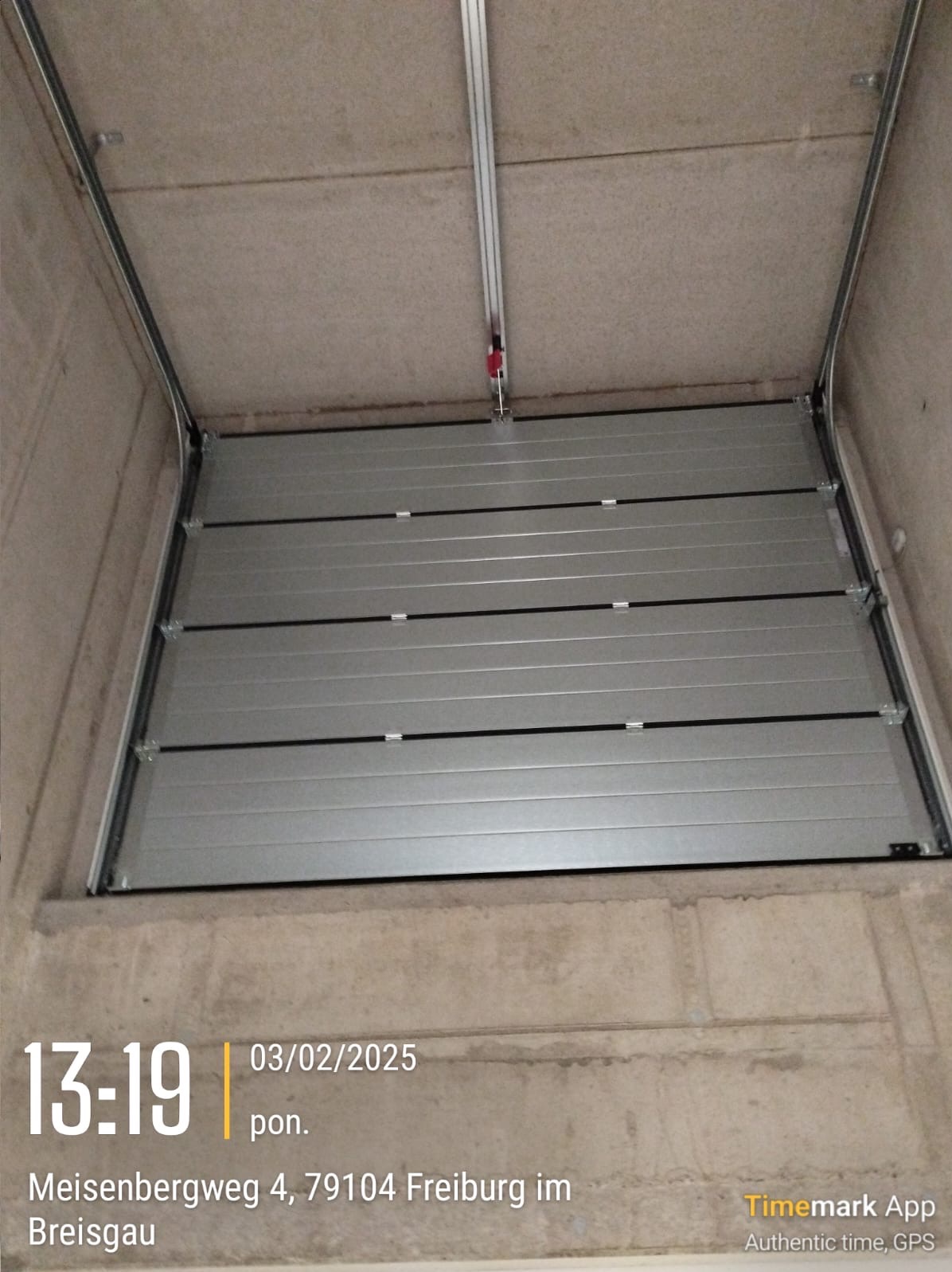 Zamknięta, segmentowa brama garażowa w kolorze srebrnym, widziana od wewnątrz garażu z widocznym mechanizmem prowadnic i betonowym sufitem oraz ścianami.