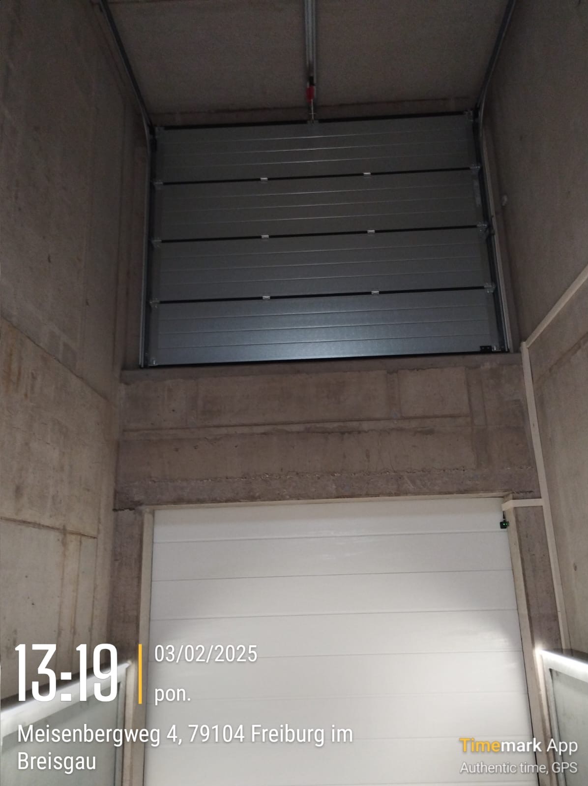 Dwie bramy w garażu podziemnym: górna segmentowa, metalowa, szara, otwarta; dolna biała, zamknięta. Widoczne surowe betonowe ściany i strop.