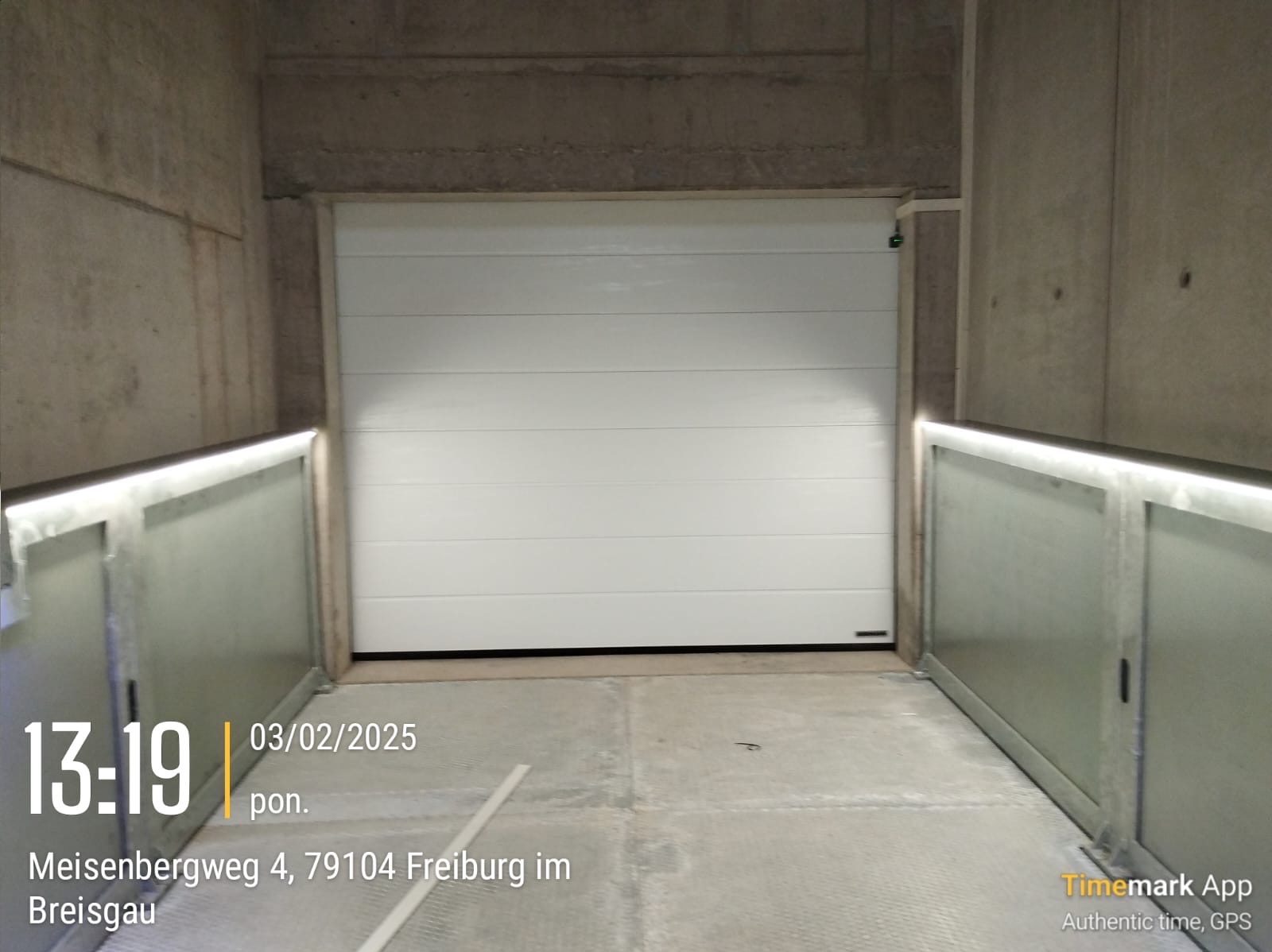 Zamknięta, segmentowa, biała brama w garażu o surowych betonowych ścianach, z dodatkowym oświetleniem LED po bokach. Widoczny znak producenta w prawym dolnym rogu bramy.