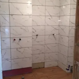 Łazienka w trakcie remontu: ściany wyłożone białymi płytkami imitującymi marmur, podłoga z paneli drewnopodobnych, widoczne instalacje wodne i notatki na ścianie.