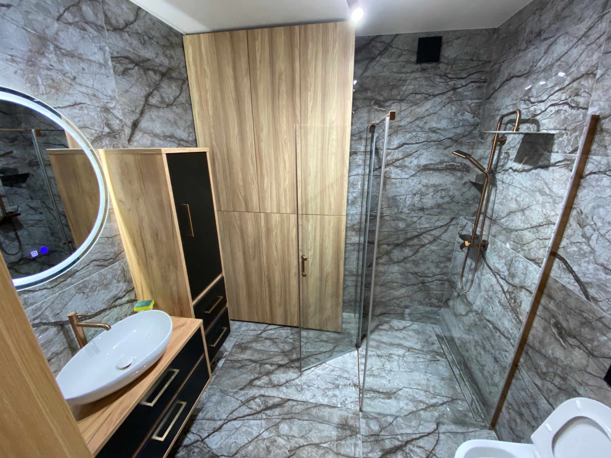 Nowoczesna łazienka z marmurowymi płytkami, drewnianymi szafkami, owalną umywalką i kabiną prysznicową ze złotą armaturą.