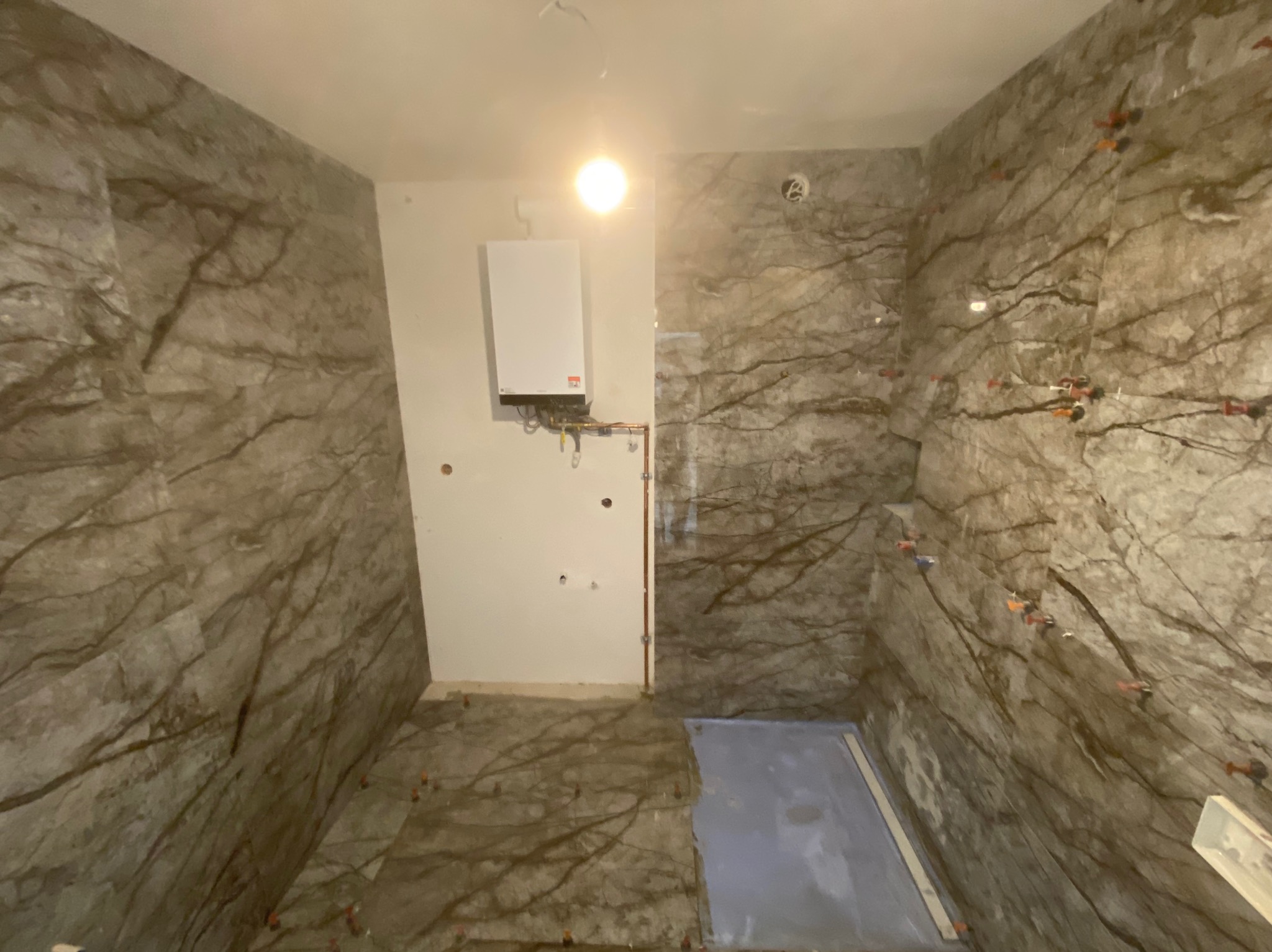 Łazienka w trakcie wykańczania, ściany i podłoga wyłożone płytkami imitującymi marmur z widocznymi krzyżykami dystansowymi, na ścianie zamontowany piec gazowy, brak fug.