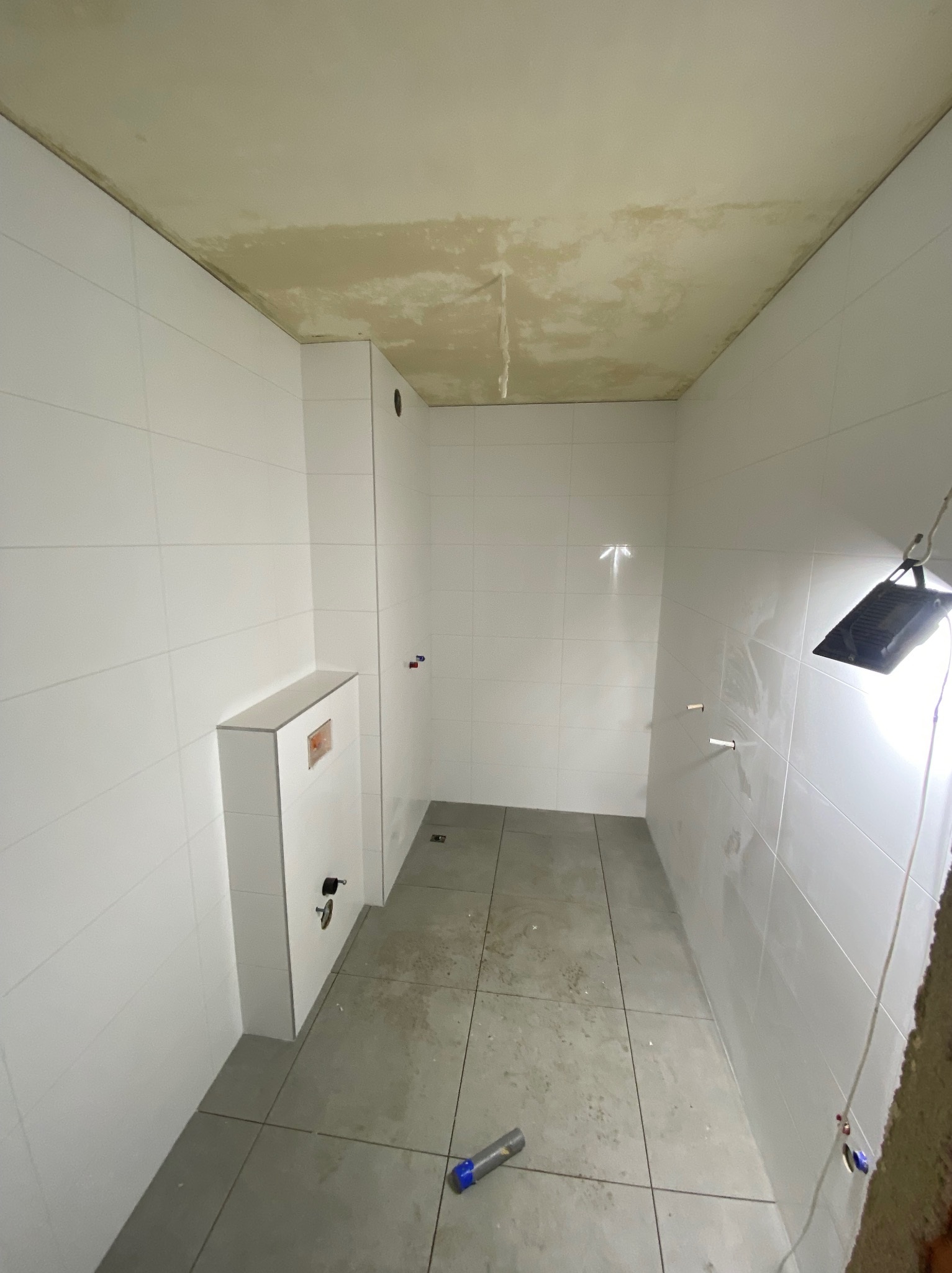 Wnętrze łazienki w trakcie remontu: białe płytki ścienne, szare płytki podłogowe, zabudowa WC, surowy sufit, lampa budowlana.