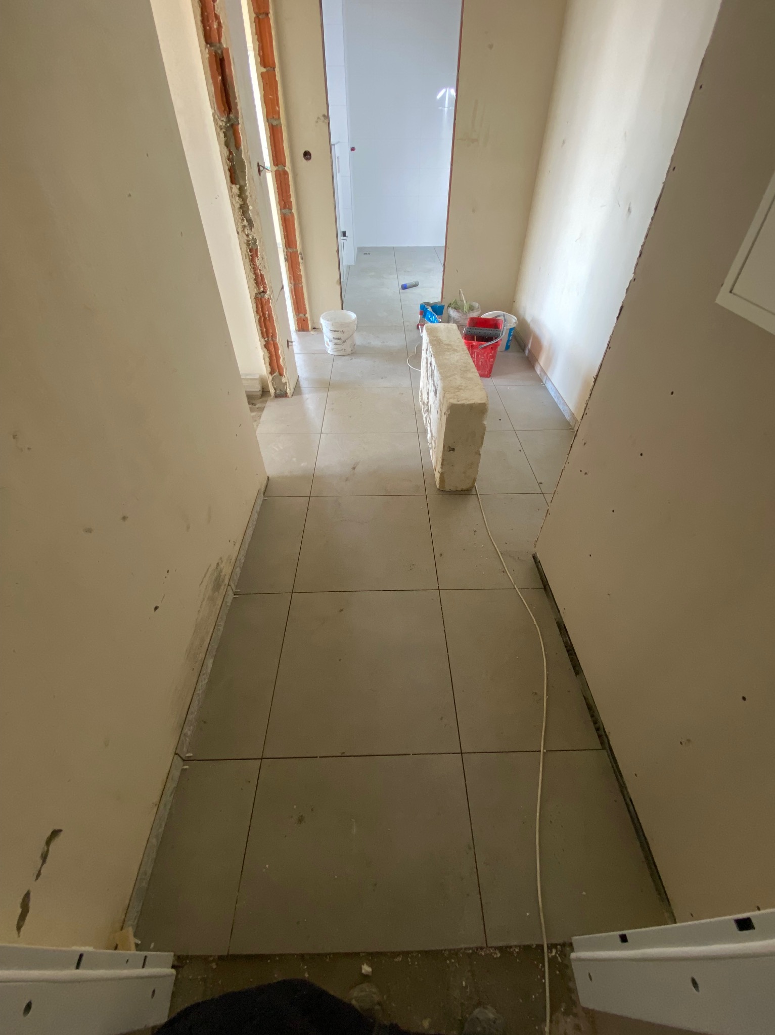 Korytarz w trakcie remontu z ułożonymi szarymi płytkami podłogowymi, widoczne puste wiadra, fragmenty cegieł i instalacja elektryczna.