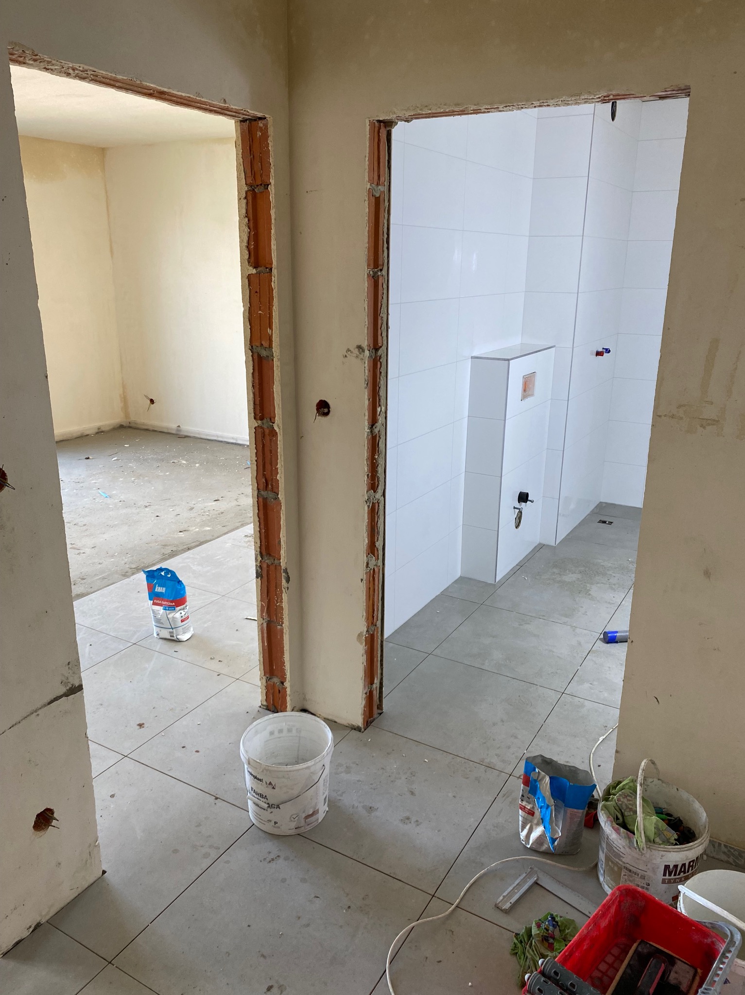 Wnętrze w trakcie remontu: widok na łazienkę z białymi płytkami i podtynkową toaletą przez surową futrynę, na podłodze płytki, wiadro z farbą i worek z fugą.