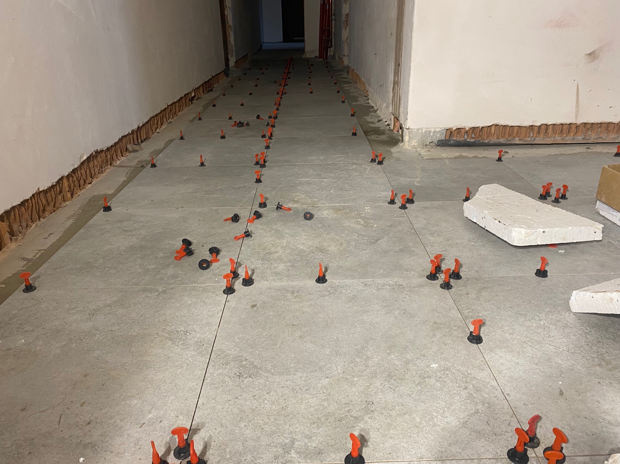 Szare płytki podłogowe w trakcie układania, z użyciem systemu poziomowania z czarnymi podstawkami i pomarańczowymi klinami, widoczne sznurki wyznaczające linie, ściany w trakcie przygotowania.