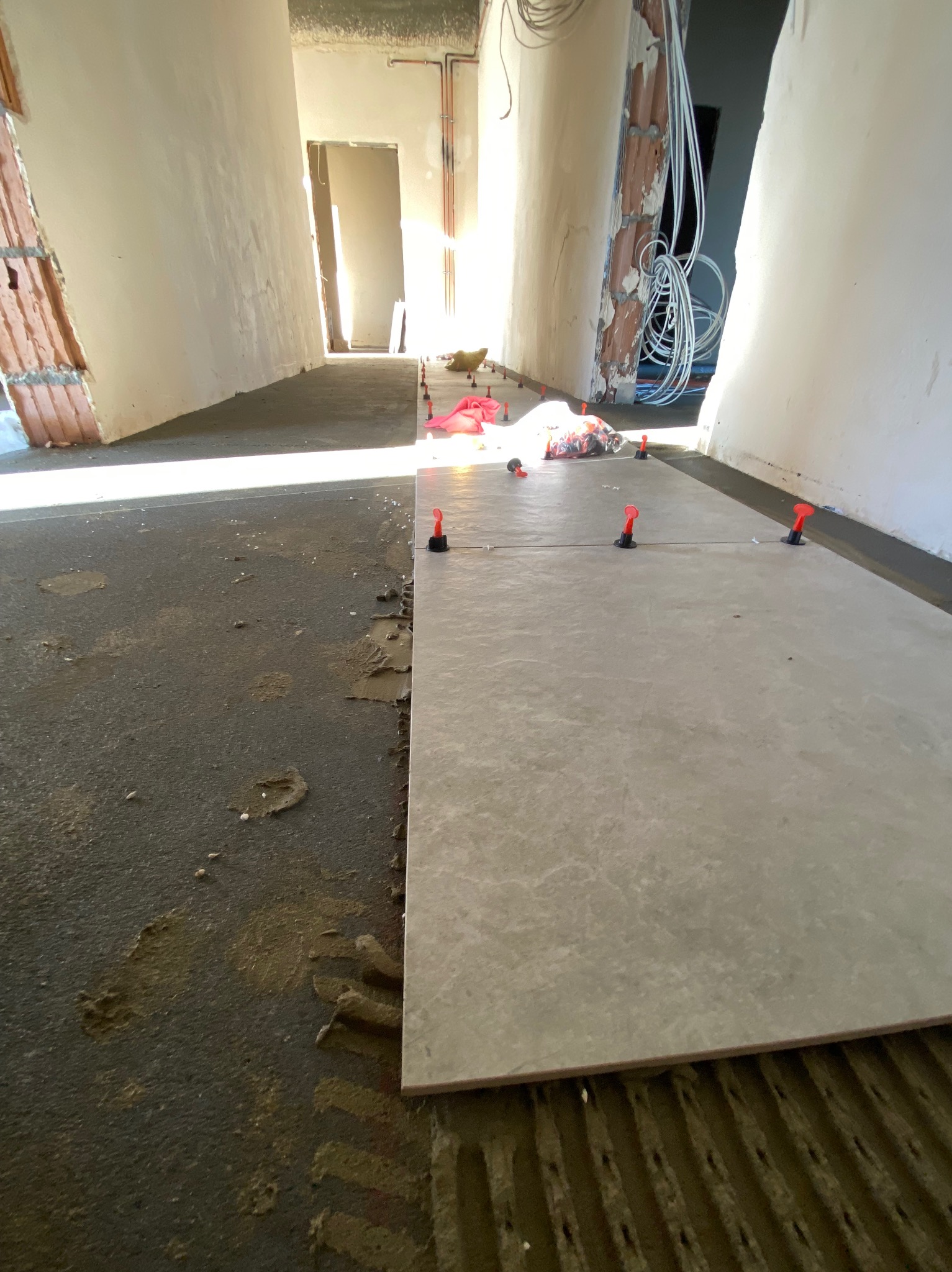 Układanie szarych płytek podłogowych w korytarzu, widoczne nierówności podłoża i system poziomowania płytek z czerwonymi klinami.