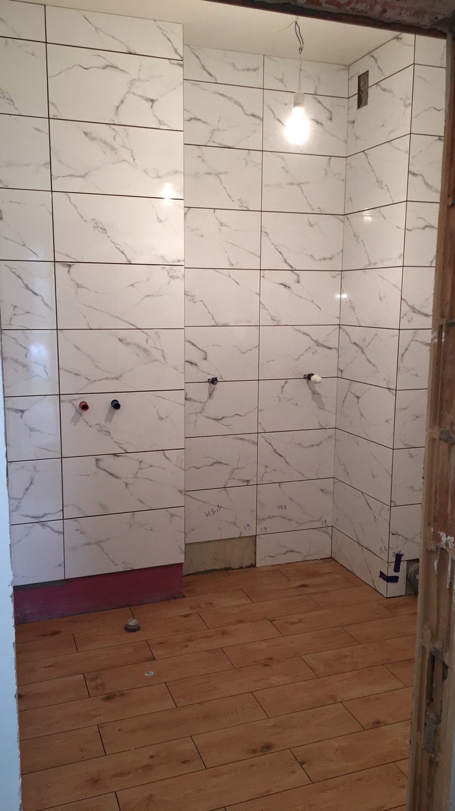 Łazienka w trakcie remontu: ściany wyłożone białymi płytkami imitującymi marmur, podłoga z paneli drewnopodobnych, widoczne instalacje wodne i notatki na ścianie.