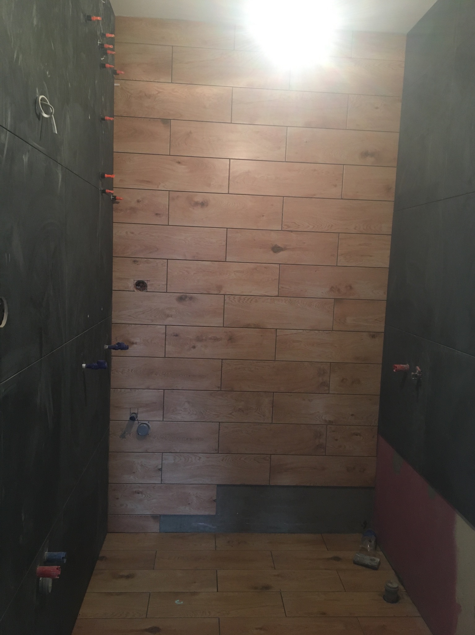 Łazienka w trakcie wykańczania: ściany częściowo wyłożone płytkami imitującymi drewno, druga ściana z ciemnych, grafitowych płytek, widoczne otwory na armaturę i poziomica na podłodze.