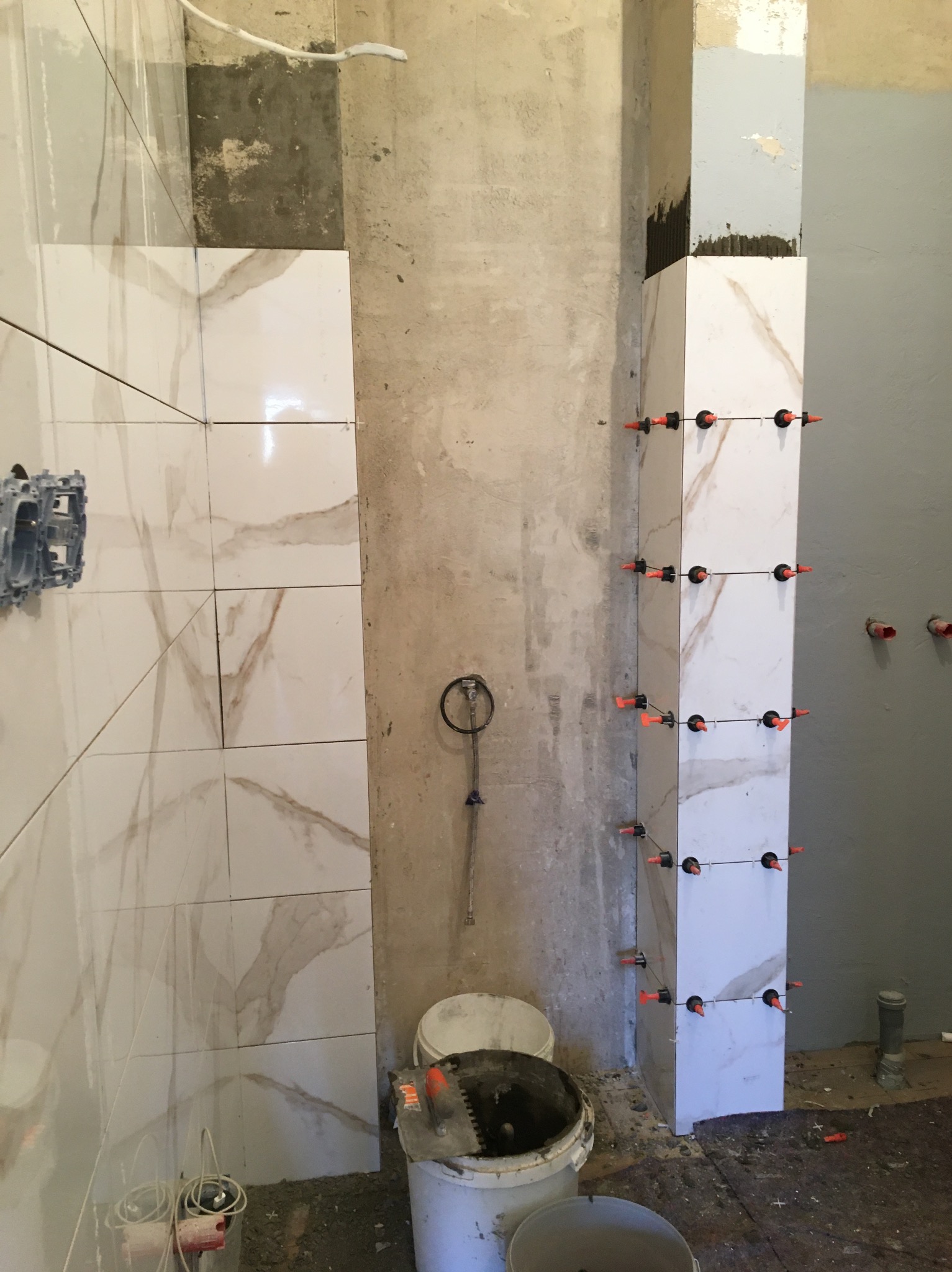 Montaż płytek imitujących marmur na ścianie i filarze w łazience, widoczne narzędzia glazurnicze i system poziomowania płytek.