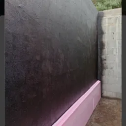 Zabezpieczona czarną powłoką i różowym styropianem ściana fundamentowa w trakcie izolacji, widoczne bloczki betonowe narożnika.