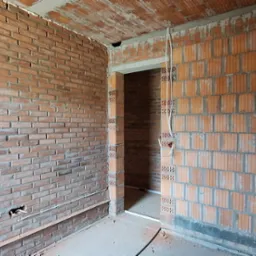Surowy mur z czerwonej cegły z widocznymi przewodami elektrycznymi w nowo budowanym pomieszczeniu. W tle widoczne wejście do innego pokoju.