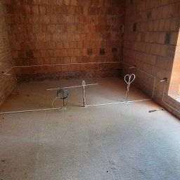 Green-Electric Ihor Kit - Surowy pokój z ceglanymi ścianami w trakcie prac. Widoczne białe rurki instalacyjne prowadzone po podłodze i ścianach oraz zwisające kable. Młotek leży na podłodze.