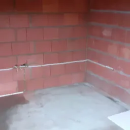 Surowe wnętrze z czerwonej cegły z widoczną białą instalacją elektryczną, kable prowadzone wzdłuż ścian. W tle widoczne inne pomieszczenia.