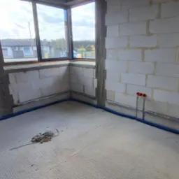 Surowy stan wnętrza z instalacją elektryczną w peszlu przy ścianach z betonu komórkowego. Widoczne puszki instalacyjne i okno z widokiem na dom i drzewa.