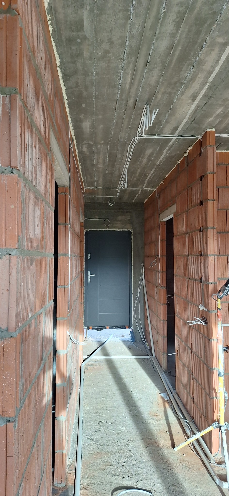 Wnętrze w trakcie budowy: surowy betonowy strop z widocznymi kablami elektrycznymi, ściany z czerwonej cegły z instalacjami, szara metalowa futryna drzwi.