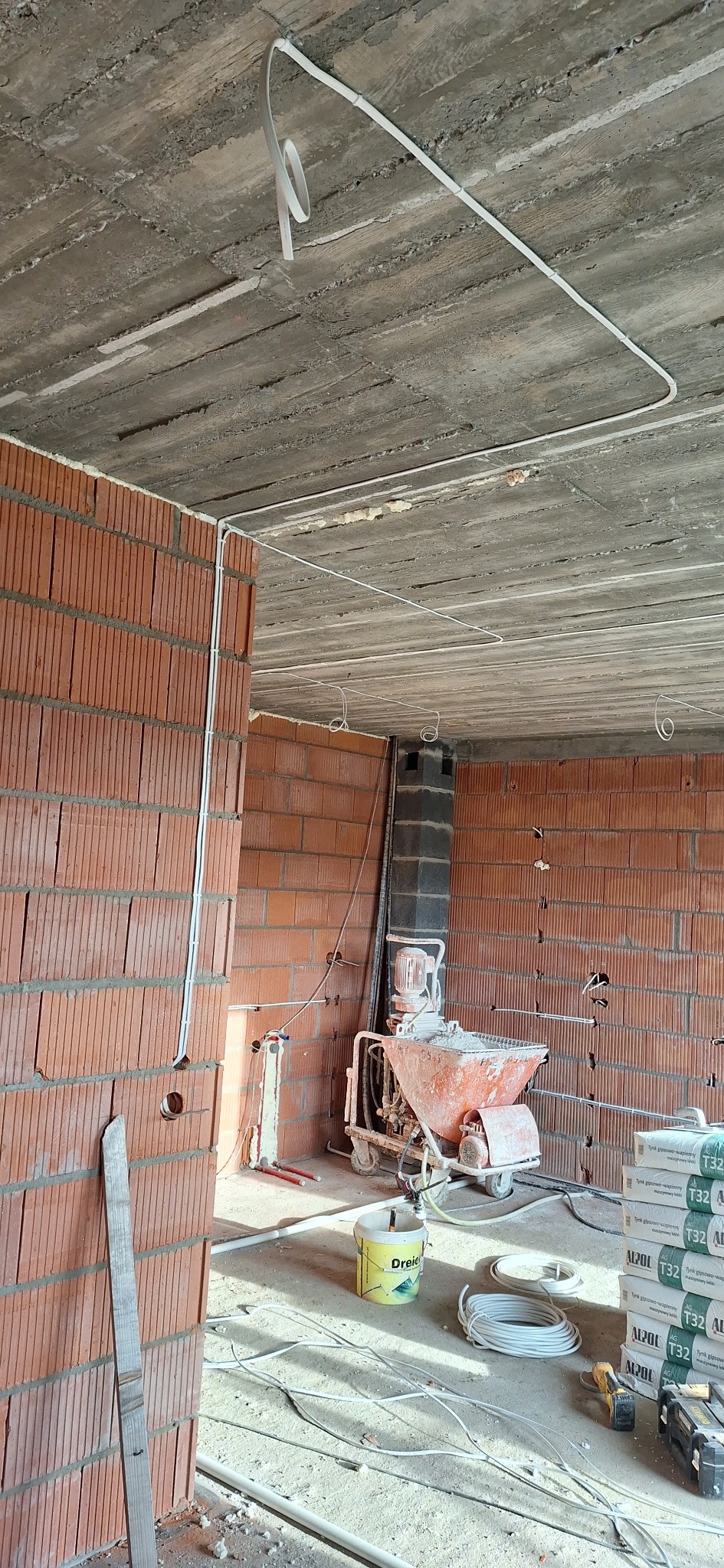 Wnętrze w stanie surowym z widoczną instalacją elektryczną na suficie i ścianach z cegły. Na podłodze leżą kable, narzędzia i maszyna do tynkowania.