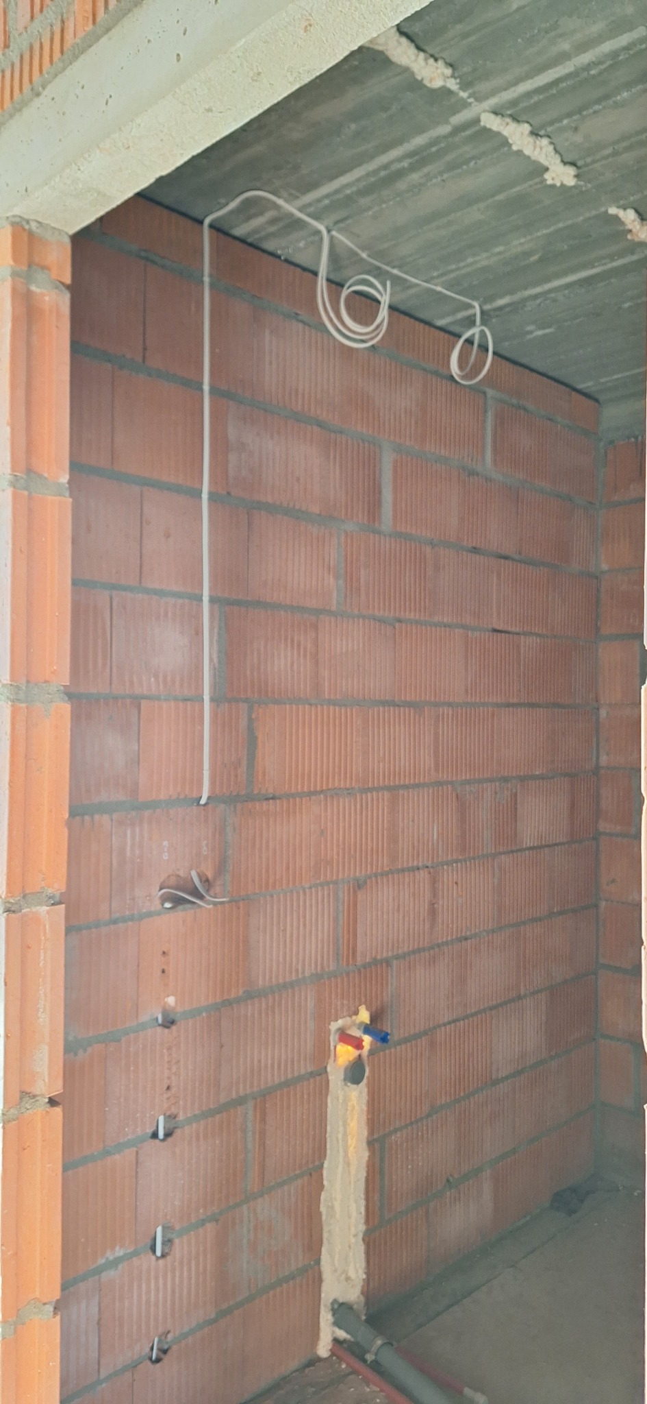 Surowa ściana z cegły z widocznymi przewodami elektrycznymi i instalacją wodną w trakcie montażu, wnętrze budynku w stanie deweloperskim.