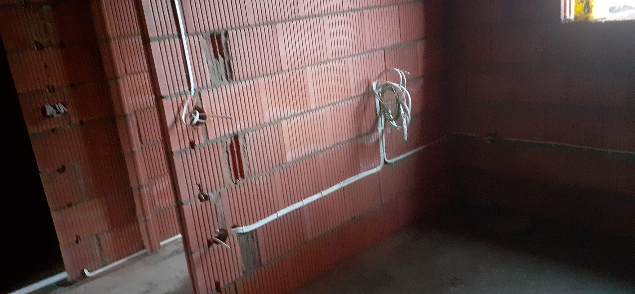 Wnętrze w trakcie budowy z widocznymi białymi przewodami elektrycznymi, częściowo zamontowanymi w czerwonej ceglanej ścianie z wyżłobieniami.