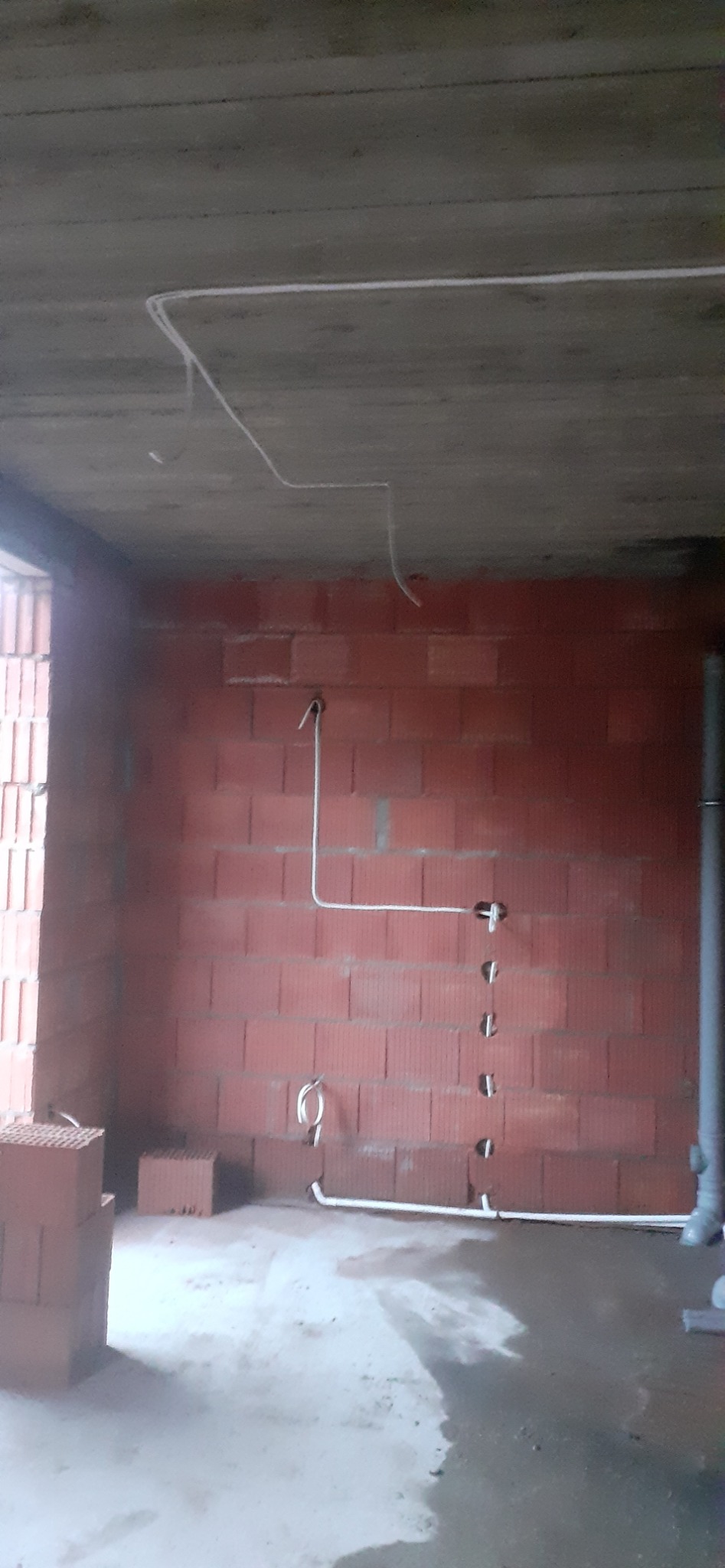Wnętrze w stanie surowym z widoczną instalacją elektryczną: białe przewody poprowadzone po ścianach z czerwonej cegły i suficie. Widoczne otwory pod gniazdka.