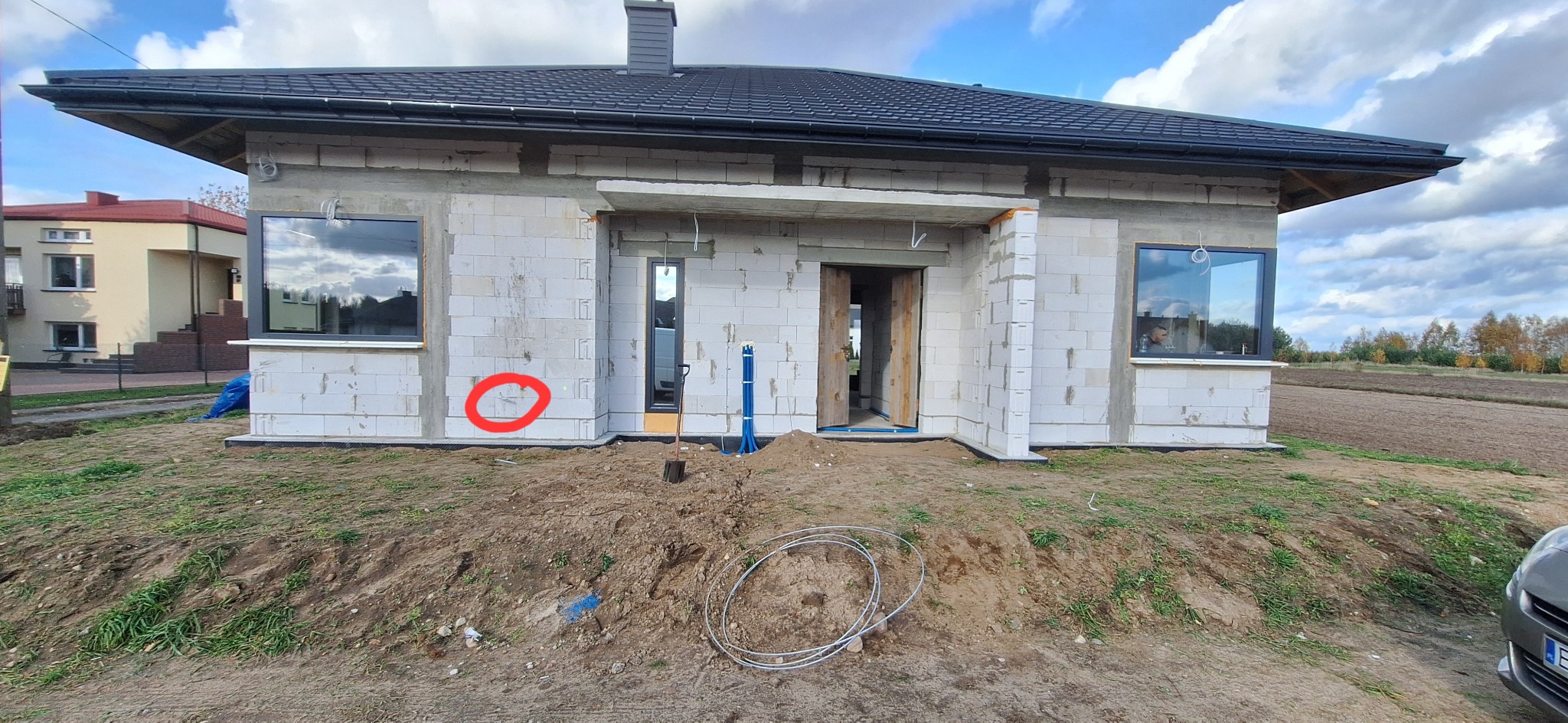 Surowy stan domu z betonu komórkowego, widoczne okna, instalacja elektryczna w ścianie, kable na ziemi. Budowa w toku, krajobraz polny w tle.