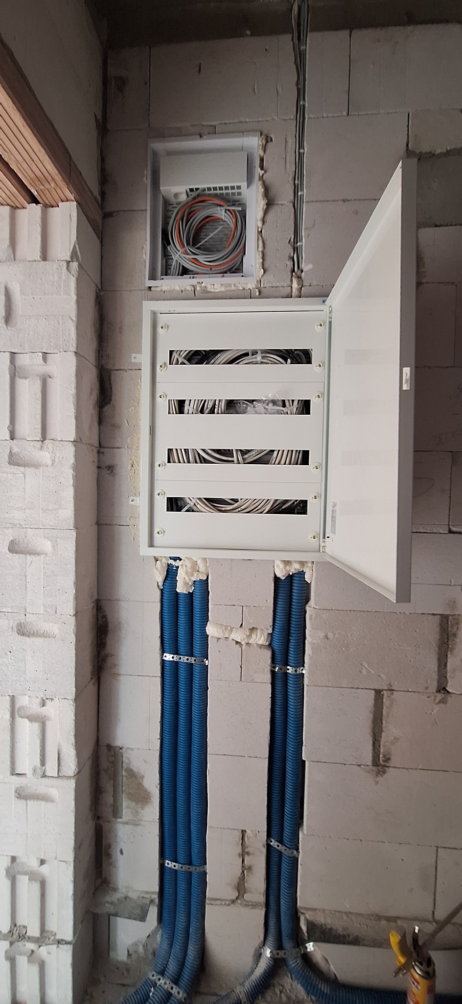 Otwarta skrzynka elektryczna z kablami w surowym wnętrzu. Niebieskie rury instalacyjne prowadzą w dół ściany z betonu komórkowego. Widoczne kable i pianka montażowa.