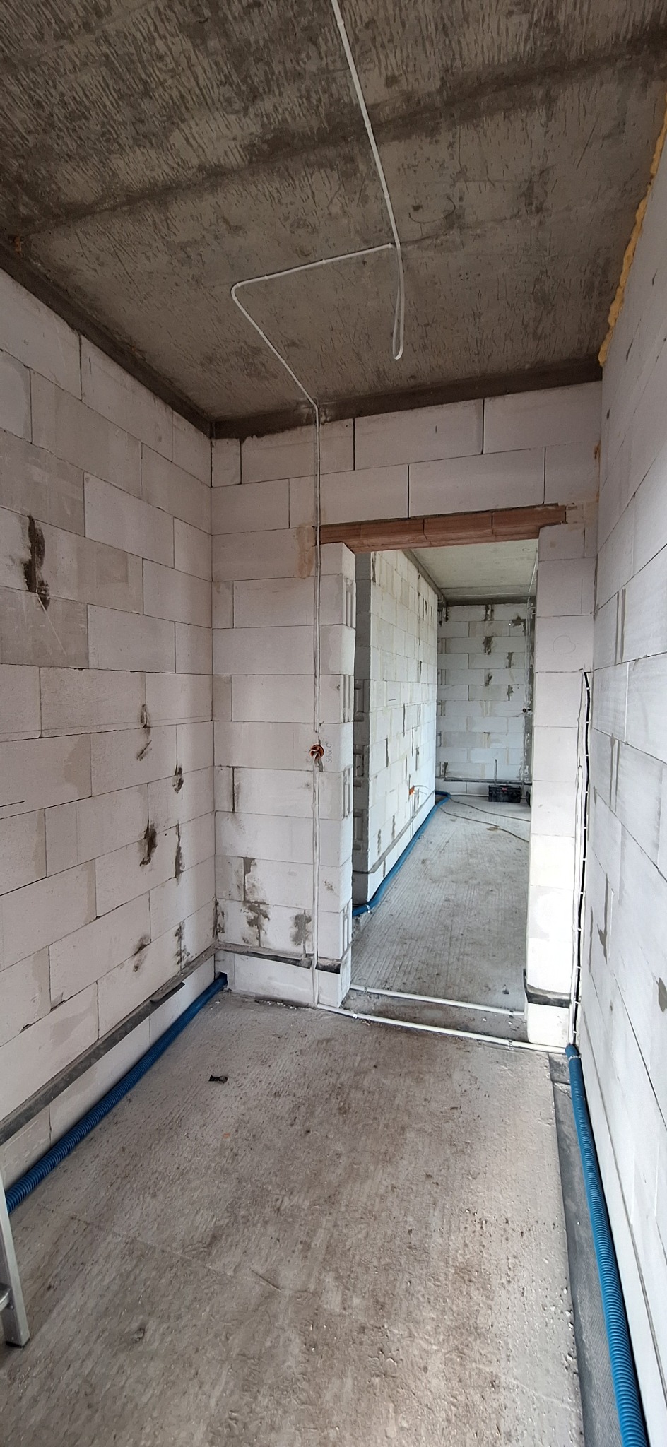 Surowy stan: wnętrze w trakcie prac z widoczną instalacją elektryczną i rurami wzdłuż ścian. Białe ściany z betonu komórkowego, surowy betonowy strop.