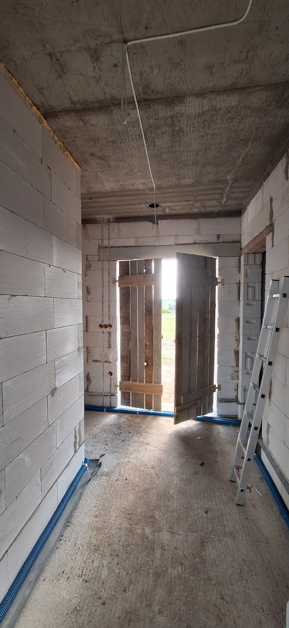 Wnętrze w stanie surowym: ściany z betonu komórkowego, widoczne przewody elektryczne, rury instalacyjne wzdłuż ścian, drabina oparta o ścianę, drewniane drzwi na zewnątrz.