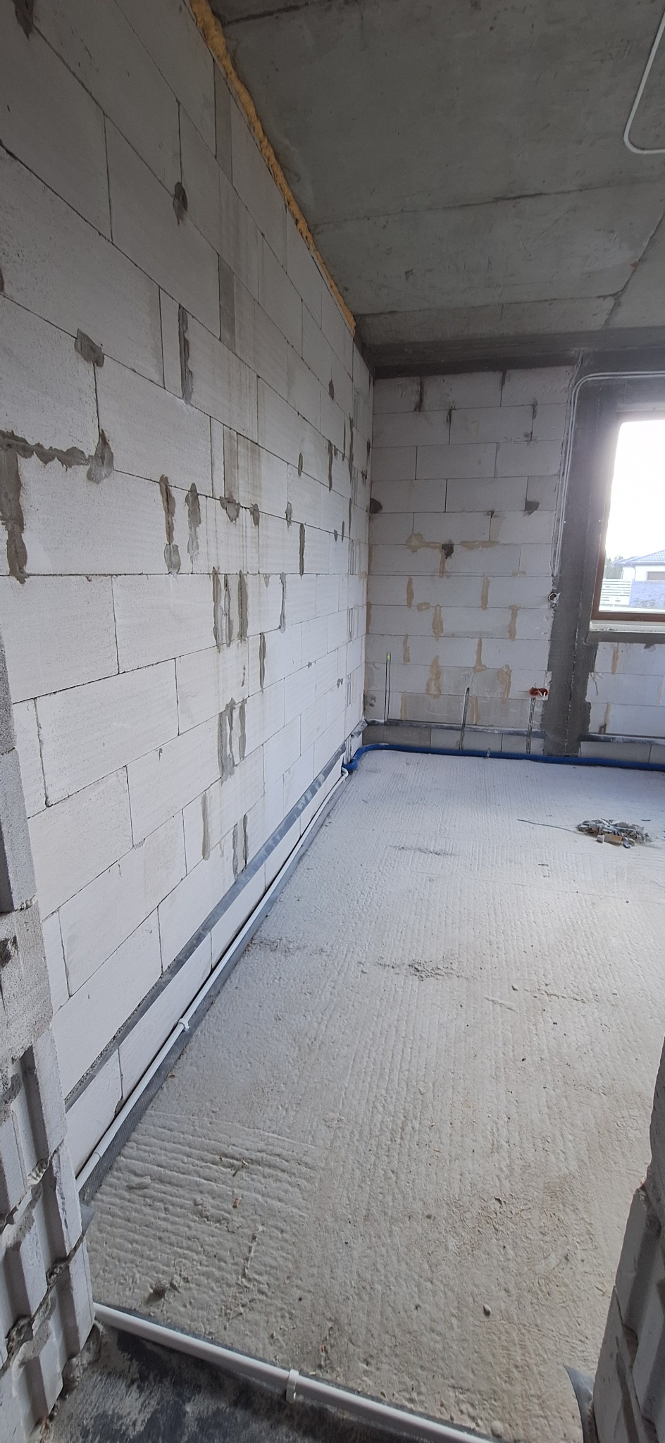Surowy stan wnętrza z instalacją elektryczną w listwach przypodłogowych i rurami. Ściany z białego betonu komórkowego, surowy betonowy strop, okno w tle.