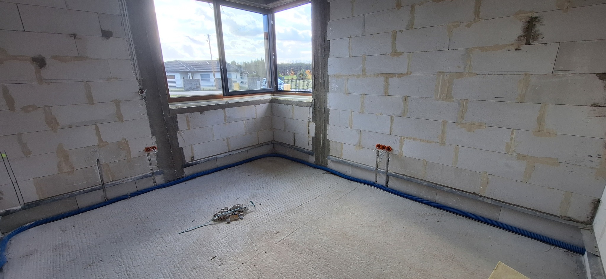 Surowy stan wnętrza z instalacją elektryczną w peszlu przy ścianach z betonu komórkowego. Widoczne puszki instalacyjne i okno z widokiem na dom i drzewa.