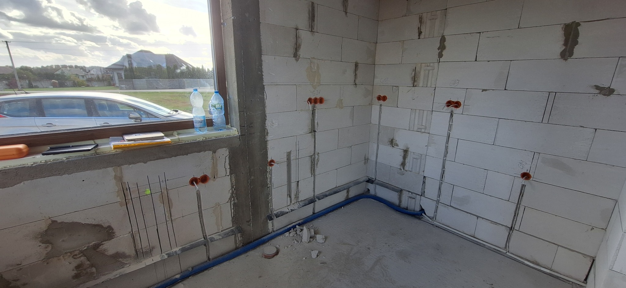 Surowy stan: ściany z betonu komórkowego z widoczną instalacją elektryczną w peszlach i puszkach, okno z widokiem na dom i samochód. Wnętrze w trakcie budowy.