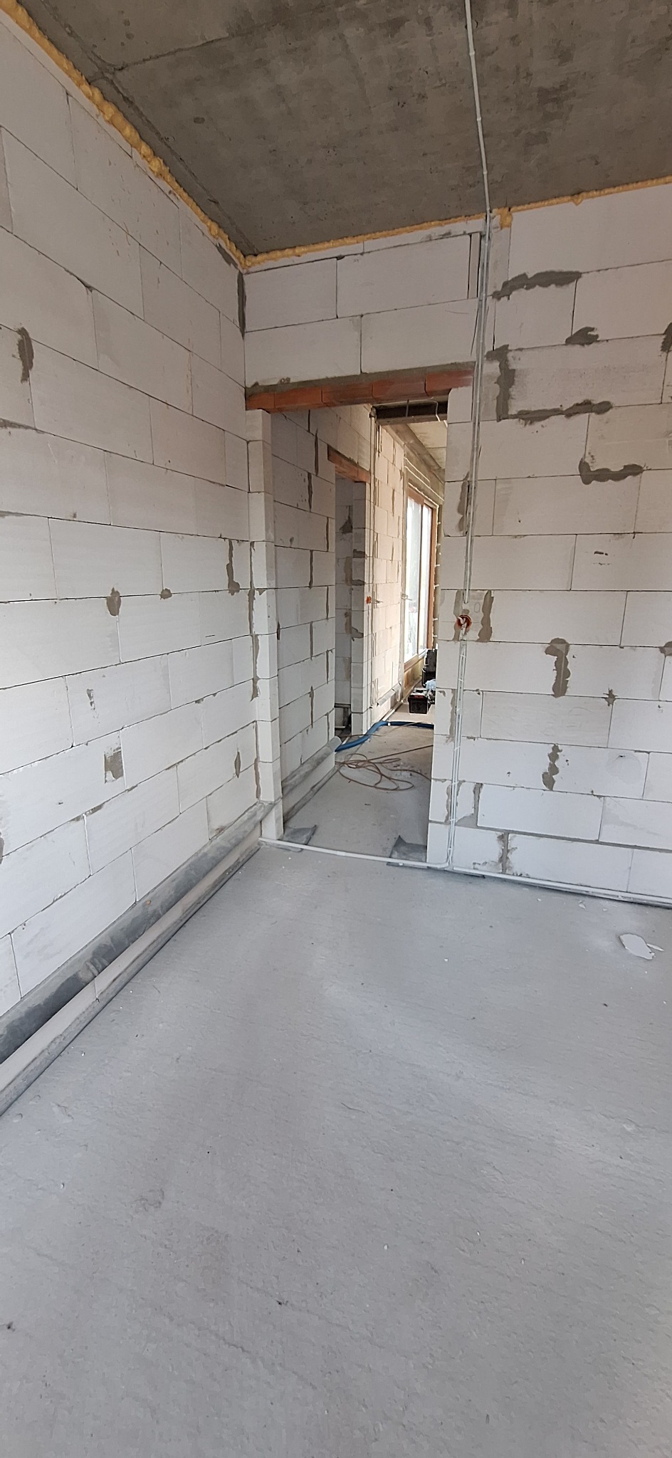 Surowy stan wnętrza budynku w trakcie prac: widoczne białe bloczki, instalacje elektryczne w peszlach na ścianie, pianka montażowa pod sufitem, surowa wylewka.