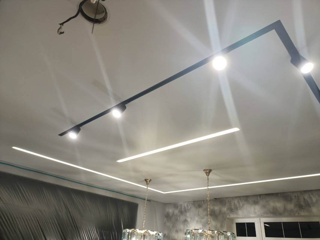 Biały sufit z zamontowanym czarnym systemem szynowym oświetleniowym z czterema reflektorami punktowymi, liniowym oświetleniem LED oraz dwoma kryształowymi żyrandolami w tle szara tapeta z motywem...