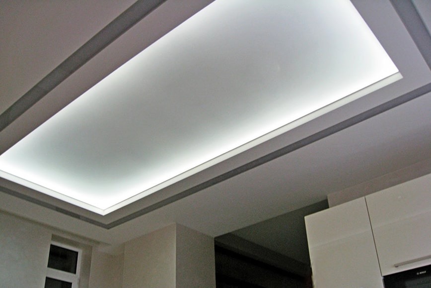 Prostokątna, podświetlana konstrukcja sufitowa z szarymi listwami dekoracyjnymi w nowoczesnym wnętrzu, widoczna część okna i zabudowy kuchennej.