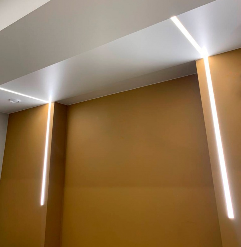 Nowoczesne oświetlenie liniowe LED w kolorze białym, wkomponowane w sufit i ścianę o barwie musztardowej, tworzące minimalistyczny design wnętrza.