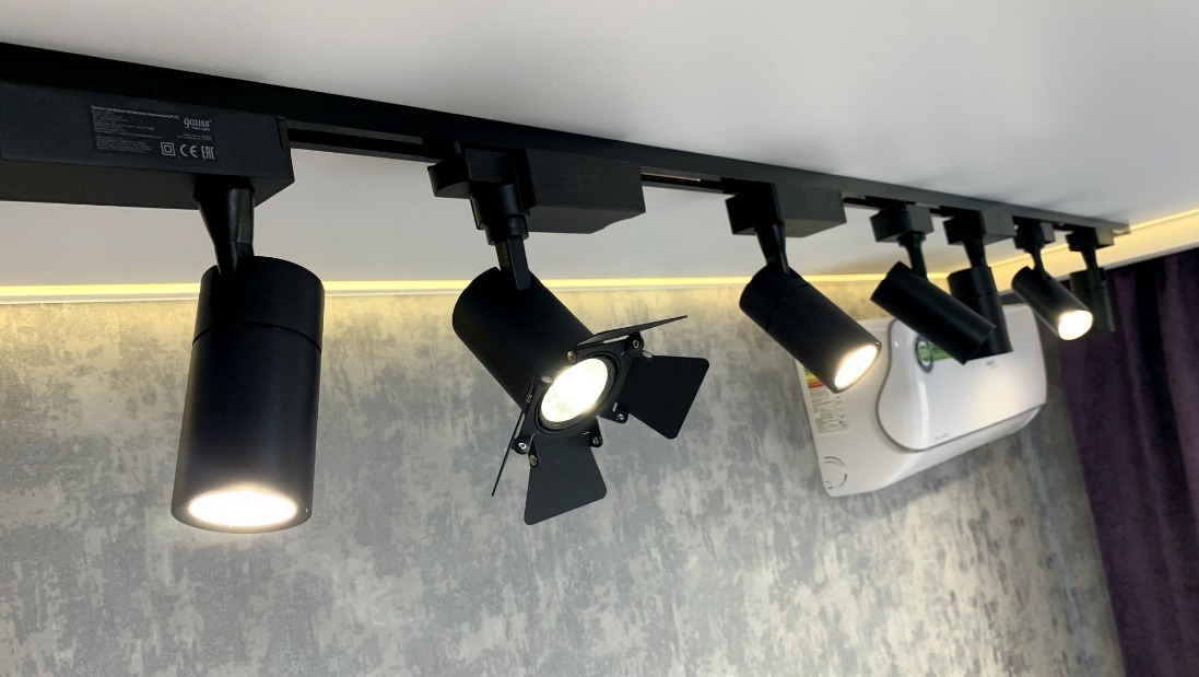 Czarny system szynowy z reflektorami punktowymi, w tym jeden z klapkami, zamontowany na suficie, widoczny klimatyzator na ścianie.