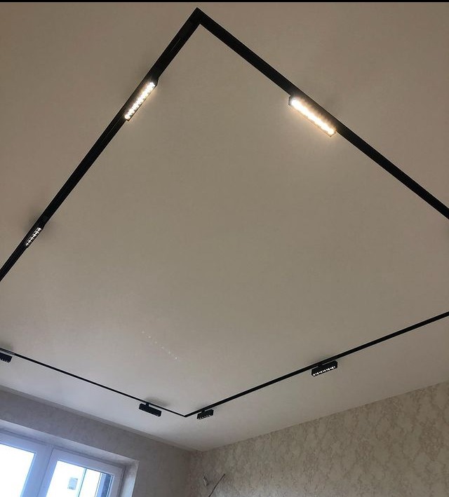 Biały sufit z czarnym, kwadratowym systemem szynowym oświetlenia LED z reflektorami punktowymi i liniowymi listwami świetlnymi.