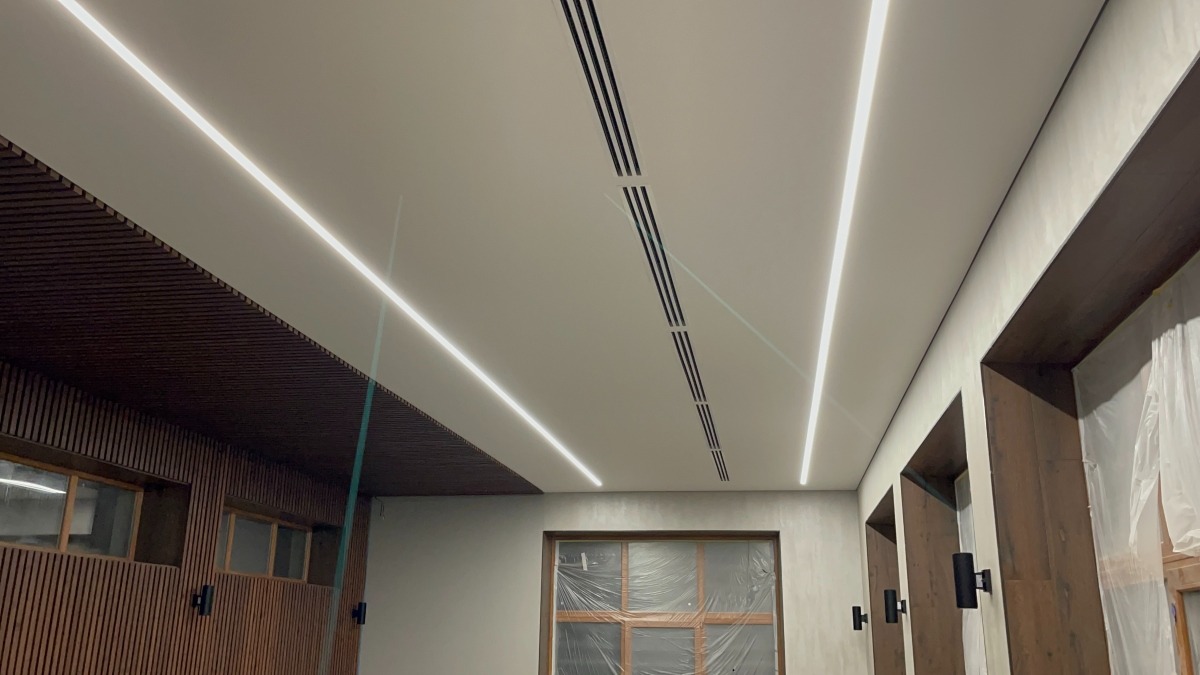 Nowoczesny sufit napinany z wbudowanym oświetleniem liniowym LED i czarnymi kratkami wentylacyjnymi, widoczne drewniane panele ścienne i stolarka okienna zabezpieczona folią malarską.