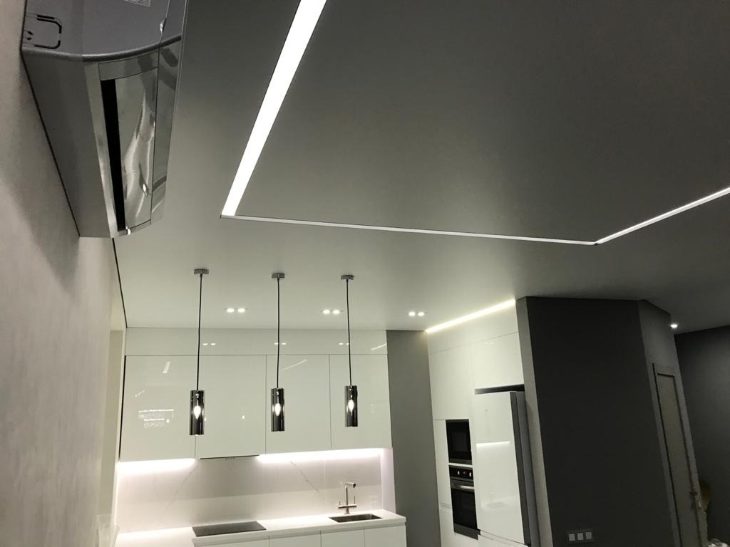 Nowoczesna kuchnia z białymi, lakierowanymi szafkami, oświetleniem LED w suficie i wiszącymi lampami nad wyspą kuchenną; klimatyzator zamontowany przy suficie.